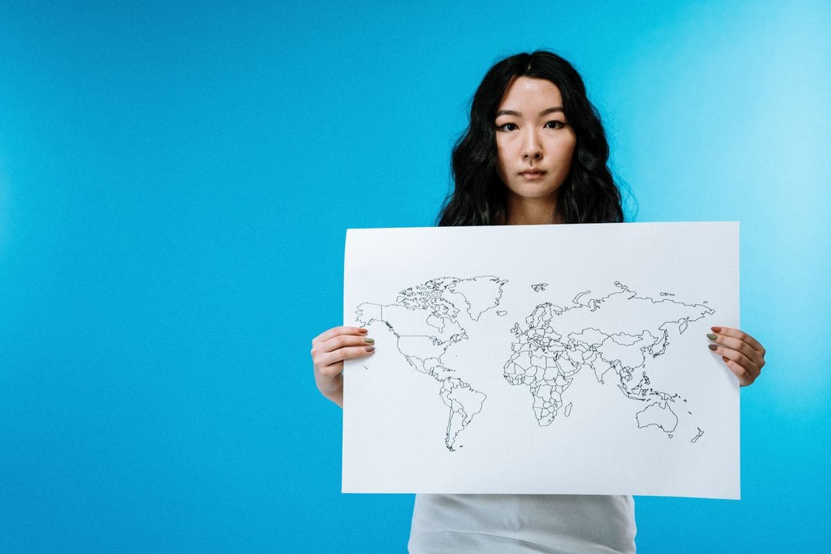 A woman with long dark hair holds up a white sheet printed with an outline map of the world against a bright blue backdrop.